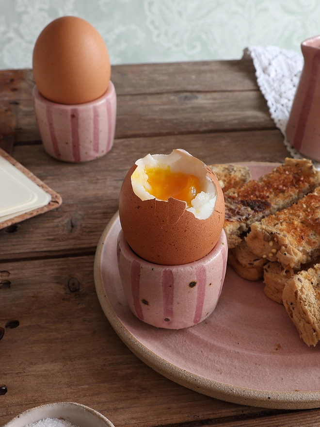 Two pink-striped ceramic egg cups holding boiled eggs, with one resting on a matching plate next to buttered toast.