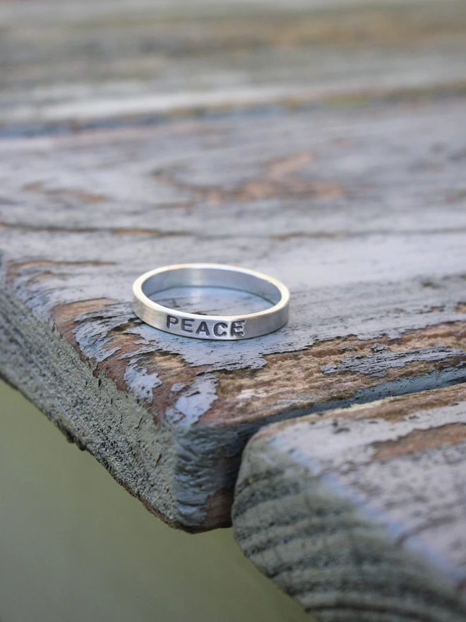 A sterling silver ring band with the word 'peace' stamped on it, resting on an aged green wooden table above a green background.