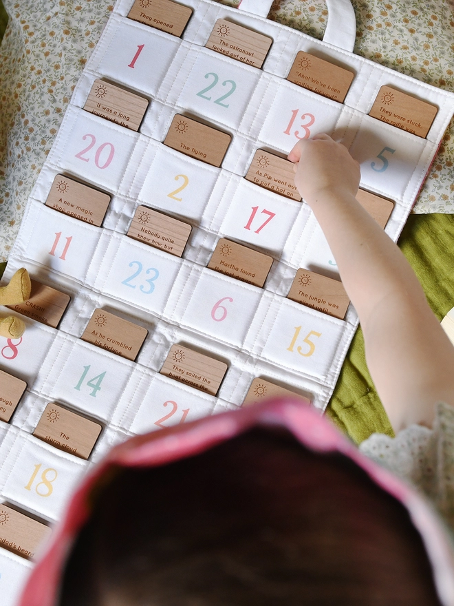 An ivory fabric advent calendar with 24 numbered pockets contains wooden cards with story ideas engraved on them. A young child reaches for one of the cards.