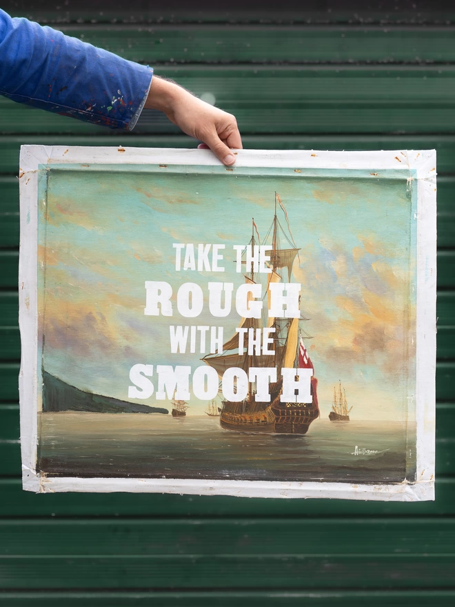 Hand holding a beautiful original canvas painting of an old sailing boat with white screen printed typography over the top with the inspirational words 'Take the rough with the smooth' 