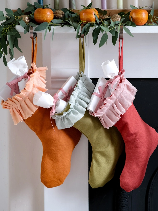 handmade linen Christmas stockings in warm orange, olive green and red hang beneath a festive mantel decorated with eucalyptus, oranges and candles, each finished with a soft, gathered ruffle cuff.