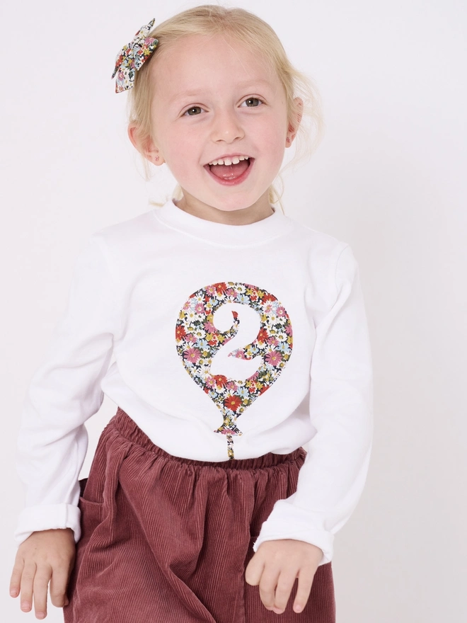 A smiling 2 year old girl wearing a white cotton long sleeve t-shirt. The t-shirt features a balloon with the number 2 cut out from it, appliquéd in a floral Liberty print.