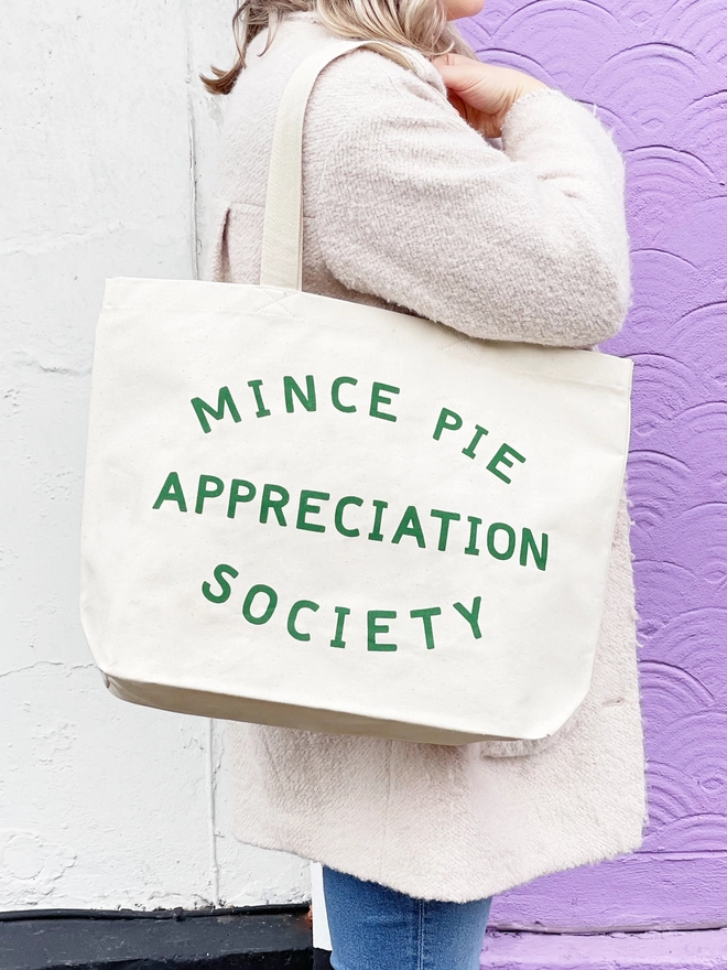 a model carrying a large natural canvas tote bag bearing the words mince pie appreciation society