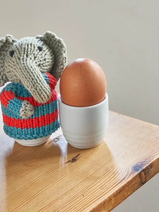 Egg Cosy Elephant Table Decoration (Set Of 2), a colourful elephant cosy sitting on a wooden surface. It is accompanied by an egg. 