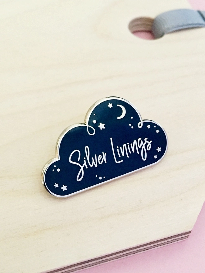 A navy blue and silver enamel pin badge in the shape of a cloud with a starry design and the words "Silver Linings" is placed on a wooden surface.