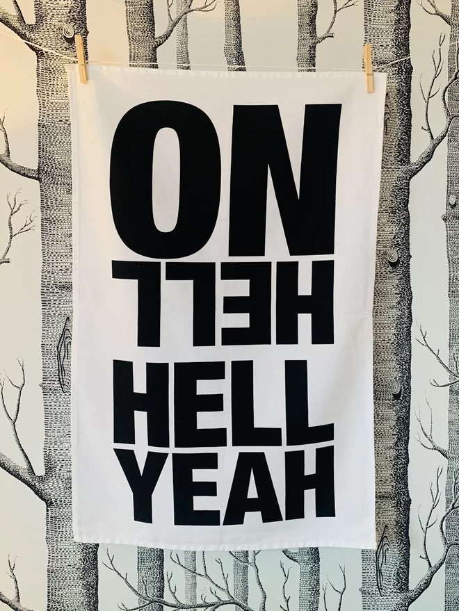 Hell Yeah Hell No black screen printed text on white tea towel hanging washing line style with clothes pegs in front of white wallpaper with black illustrated trees pattern. Hell Yeah facing forward , Hell Yeah facing backward