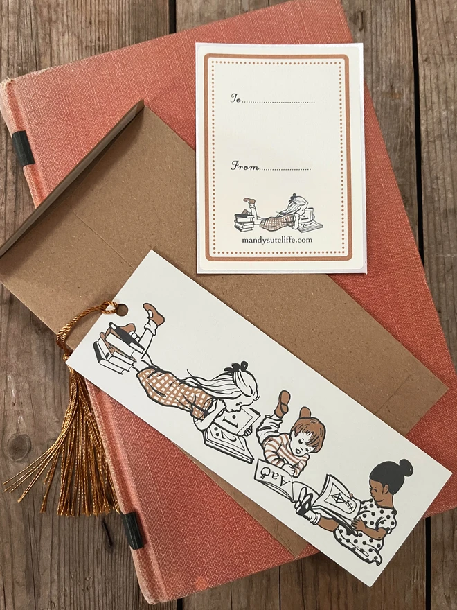 a photograph of a traditional card bookmark illustrated with a vintage children's book style drawing of kids reading books. The bookmark is cream, ochre and sepia in colour, the tassel is a golden brown. Laid on a kraft envelope and there is also a gift giving label, or bookplate sticker making it a lovely alternative to a greeting card