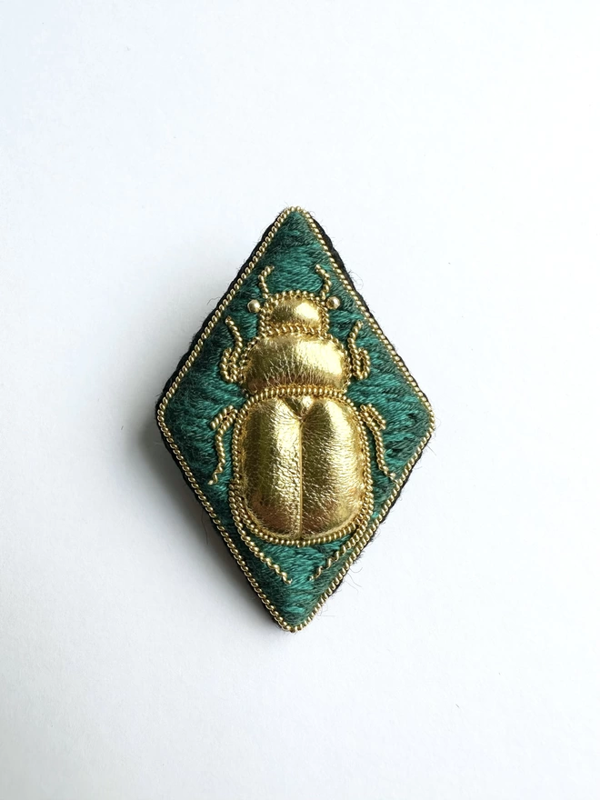Diamond shaped brooch with golden beetle on a green background