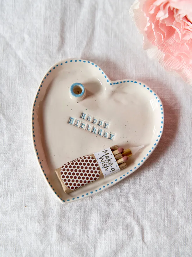 Handmade ceramic heart dish stamped with "Happy Birthday" featuring a blue dotted rim on a plain white table cloth 