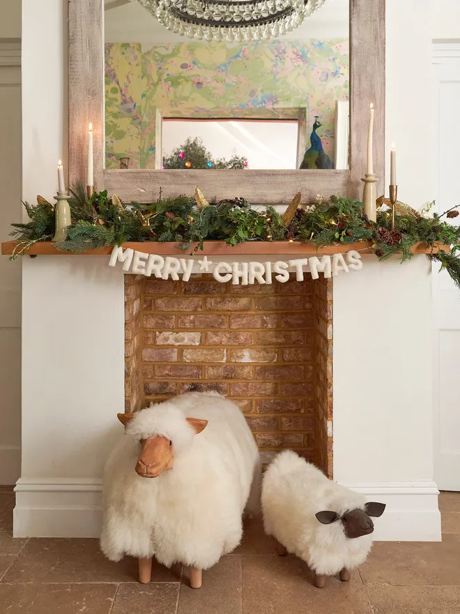 A felt Merry Christmas banner hung on a fireplace, along with foliage and candles and woolen sheep.