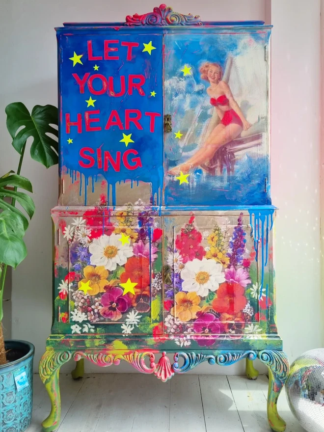 Upcycled cocktail cabinet in neons and bright colours. The decoration features a retro pin-up model on a boat and florals with stars and the wording reads ‘let your heart sing’