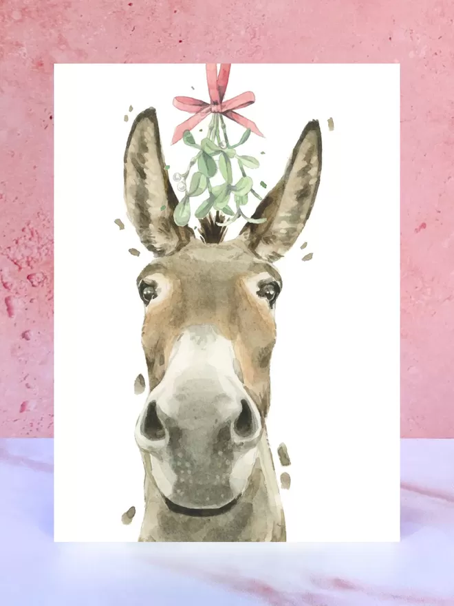 A Christmas card featuring a hand painted design of a Donkey, stood upright on a marble surface.