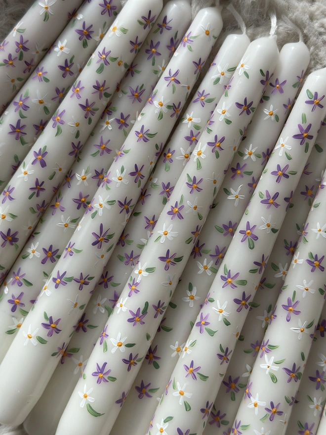 Purple Fleur Candle, a group of purple fleur candles sitting on a white rug. Their purple floral design can be seen wrapping around them. 