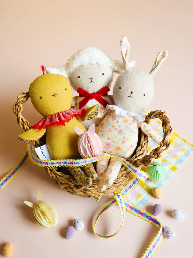 Handmade chick doll made from soft yellow fabric, with a stitched face, small beak, ruffled collar, and patterned fabric arms and long legs, with fluffy white lamb doll and floral bunny doll.