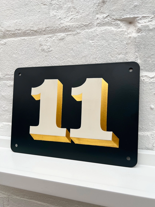 Hand painted house number against a white brick wall, number 11 in off white and directionally burnished 23 carat gold leaf, on anthracite grey background. 