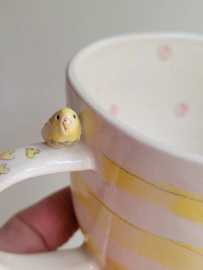 Stripey yellow and off-white cup close up with a tiny ceramic budgie perched on the side with yellow hearts