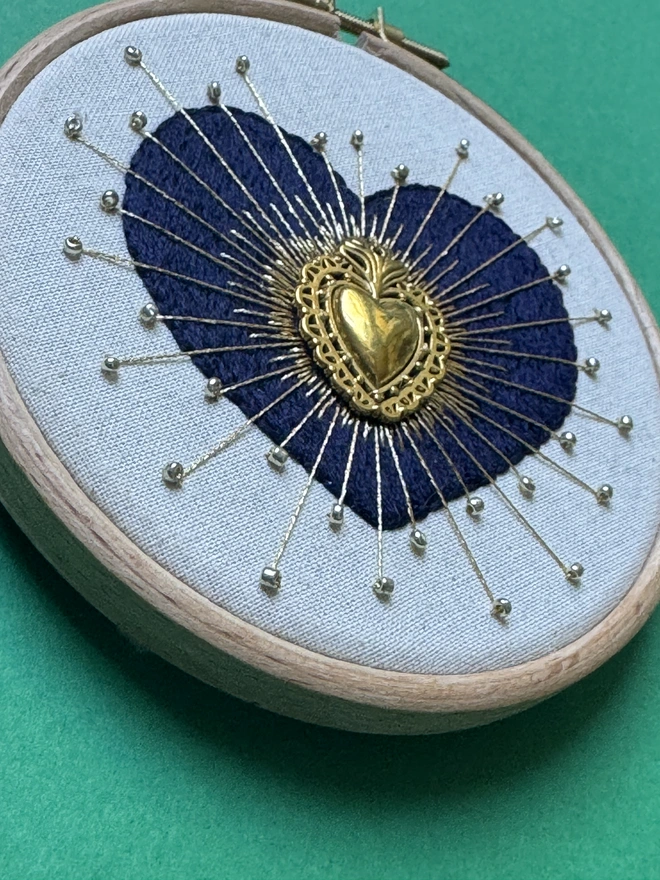 An embroidery hoop containing a hand stitched embroidered heart with gold beads and floss 