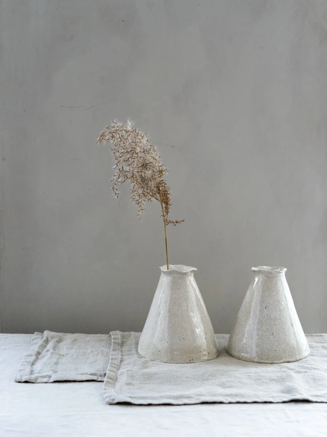 Maia Bud Vase In Sea Salt Glaze, with plant in it, placed on a table cloth against a grey background. 