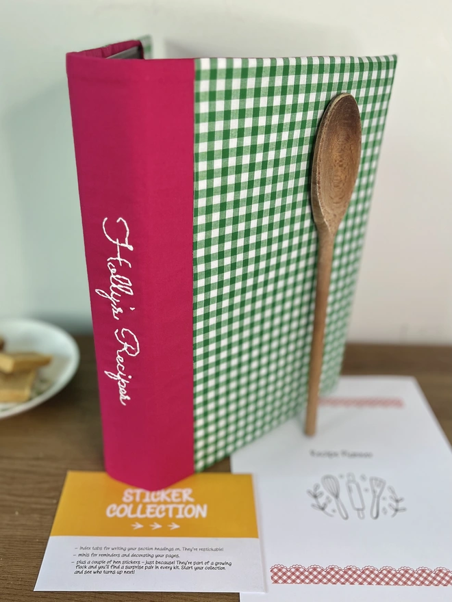 Green gingham recipe binder with 'Holly's Recipes' embroidered on pink spine, standing on wooden bench with wooden spoon propped against it and recipe template pages and 'Sticker Collection' envelope lying beside it. Plate of baking in background.