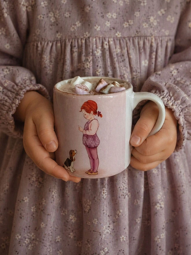a small childs ceramic mug being held in the hands of a small child featuring an illustrated mug of a little girl with red hair and her Beagle puppy teaching him to sit pink and lilac in colour