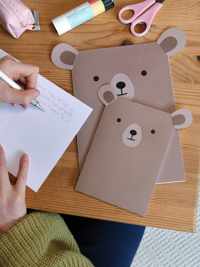 Several brown bear greetings cards lay on a wooden table. A child is writing a message in one of the cards.