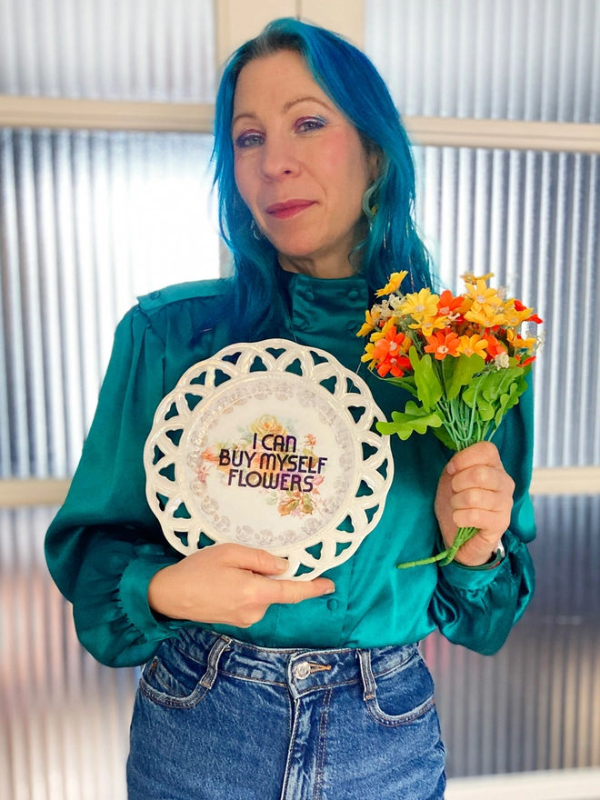 Lucy holding a plate printed with the words 'I Can Buy Myself Flowers' in her right hand and a bunch of colourful plastic flowers in her felt.