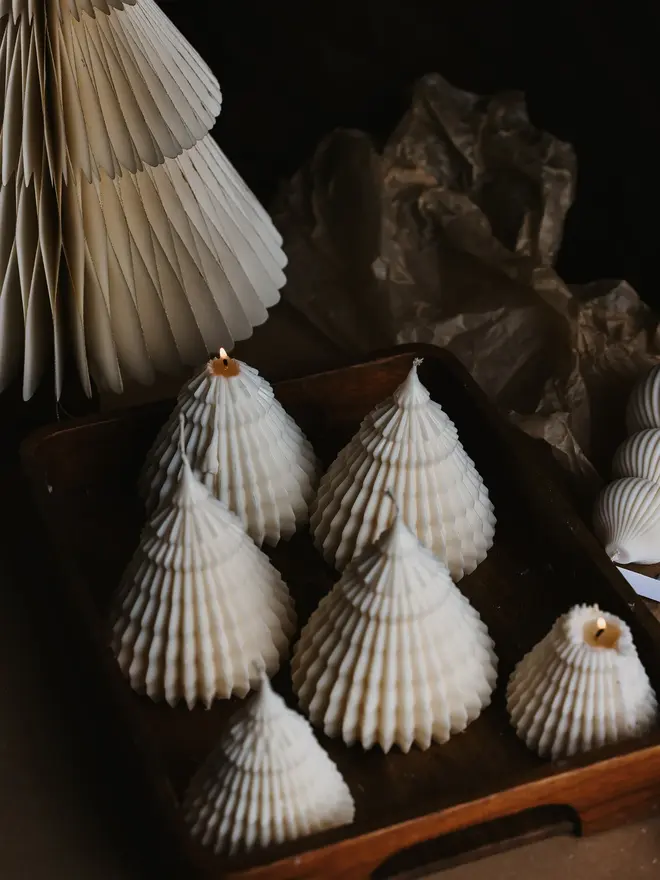 Origami Christmas Tree Candles (Set Of 2). A wooden tray filled with cream cone tree candles, two softly lit, beside a pleated paper tree and crinkled tissue paper. The sculpted texture and neutral tone suit stocking fillers, table favours and festive gift sets.