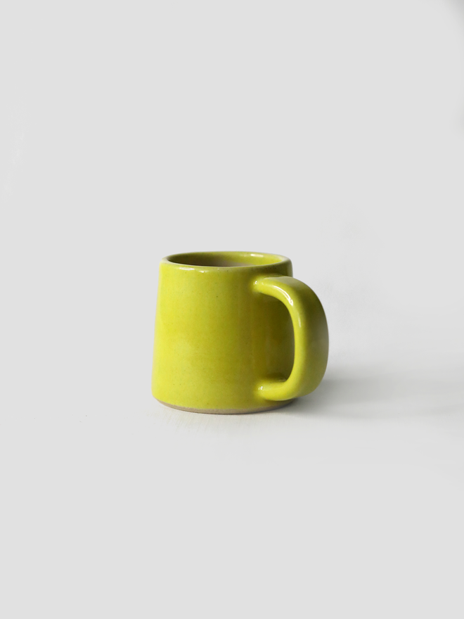 A handmade ceramic espresso mug in sun yellow glaze, with a chunky loop handle, photographed against a plain light grey background