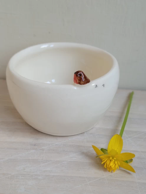 Pottery ceramic white tealight candle holder with tiny robin bird and birdprints attached on the side on a light wooden surface with a yellow buttercup flower lying next to it
