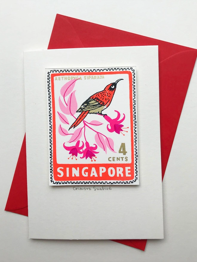 Bird Stamp Card Singapore/ Crimson Sunbird, a white card with a colourful bird design in the centre above a red envelope on top of a white surface. 