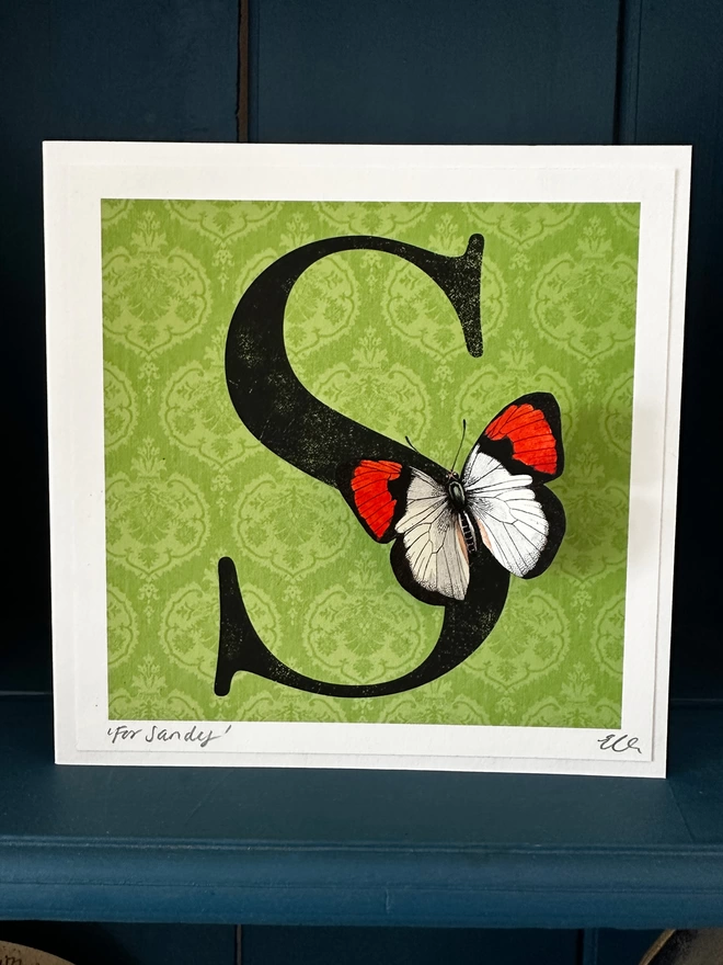 personalised birthday card with large letter on a green coloured patterned background with a 3D paper cut butterfly. Card displayed on a blue shelf in home