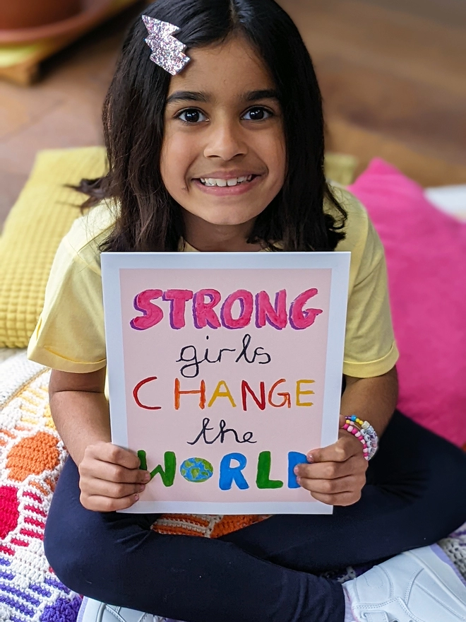 Young girl sitting holding an art print saying 'Strong girls change the world'