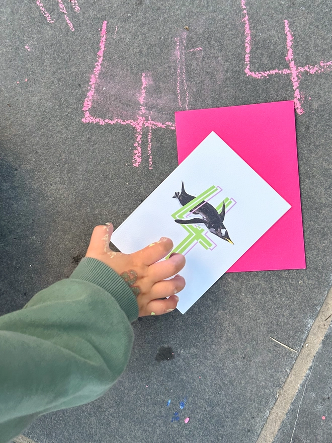 a white card with a big green number 4 and a penguin illustration with a neon pink envelope on the floor next to some chalk 4s 