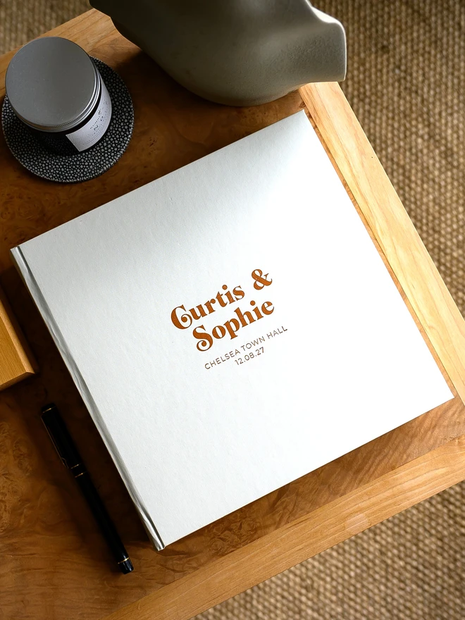 A pearl-coloured wedding guest book with gold printing on the cover rests on a wooden table.
