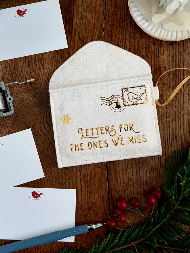 A handmade ivory felt envelope decoration with the words 'Letters for the ones we miss' in gold lays on a wooden table. Several robin notecards lay beside it.