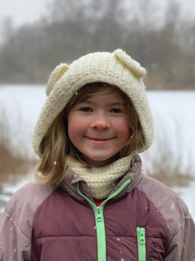 Girl smiling at camera wearing loose fitting handmade crocheted bonnet or hood  with bear ears in a cream colour.