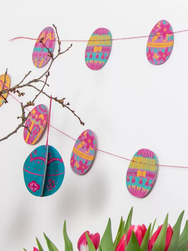 Pink, yellow and blue easter eggs hanging on white wall and indoor plant