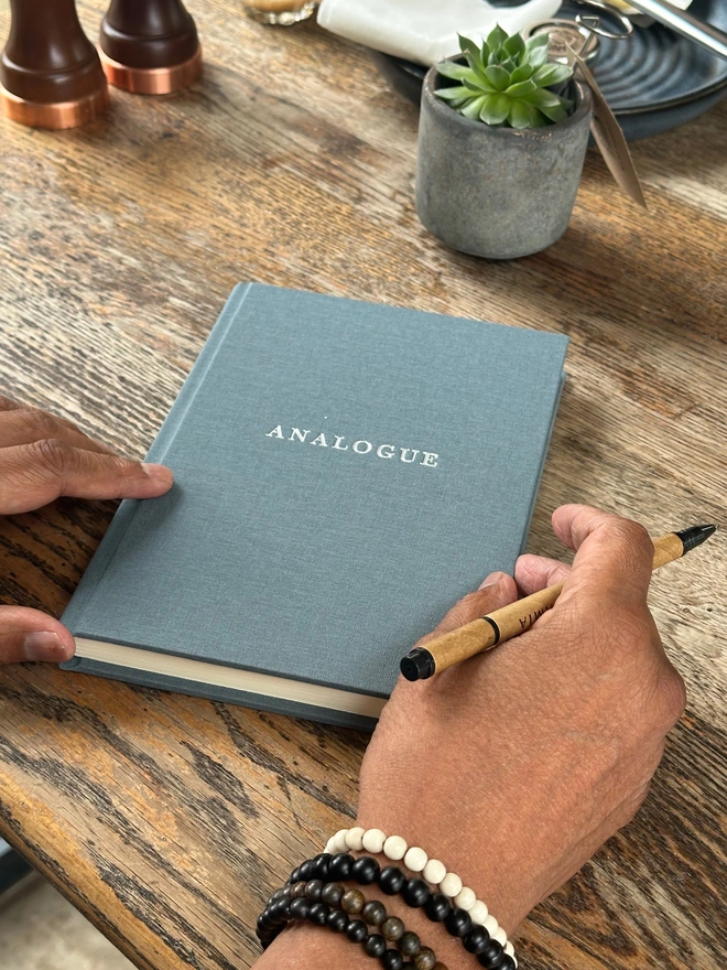 A notebook covered in grey linen and printed with 'Analogue' in gold lettering is held in a man's hands, along with a pen.