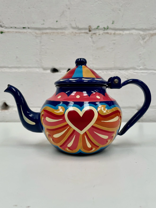 Small blue vintage enamel tea pot, hand painted with a fairground style design in pink, orange, and red. Teapot has a hinged lid.