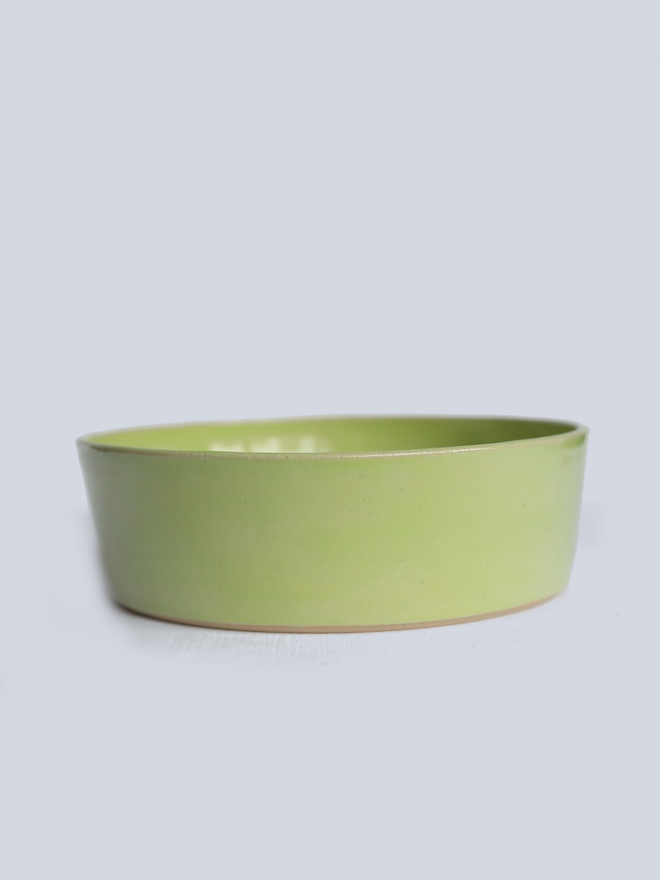 A single wide, shallow glazed ceramic pasta bowl in spring green, photographed from a slight angle against a plain light grey background, showing the unglazed natural clay base.