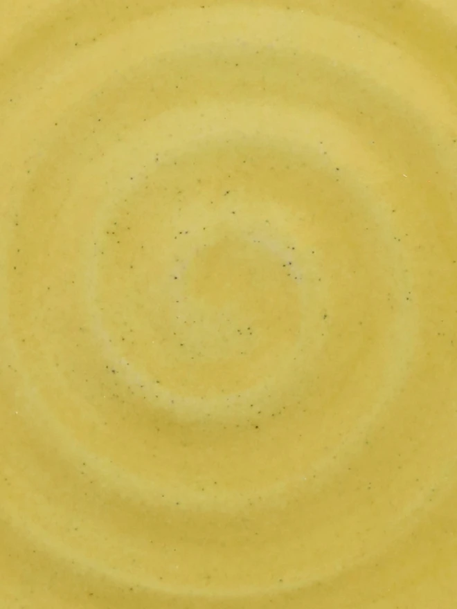 A close-up of a handmade ceramic glaze swatch in sun yellow, showing a smooth concentric swirl texture with a delicate, even tone throughout