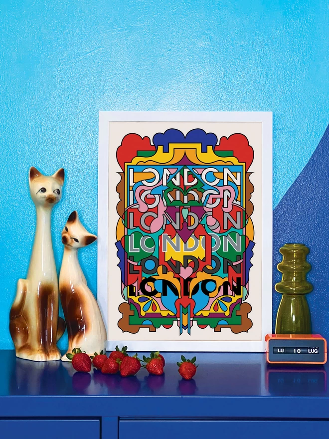A vibrant, portrait print containing all the colours of the London Underground lines, with the word London repeated six times from top to bottom. The picture is in a white frame, against a turquoise and blue wall resting on a blue cabinet. Next to the picture are two cat ornaments, some ripe strawberries, a yellow glass vase and an orange Italian plastic calendar showing the date as ‘LU 10 LUG’.