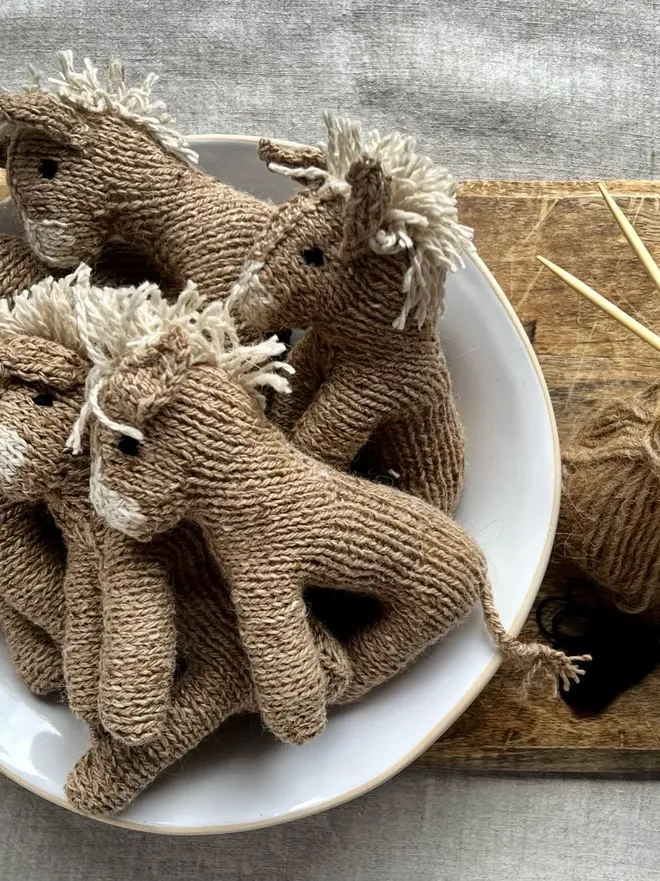 Hand Knitted Natural Dyed Wool And Alpaca Yarn Donkey Gift, a group of hand knitted donkeys sitting in a white bowl on a wooden chopping board on a table cloth. They are accompanied by some hey and some knitting accessories. 