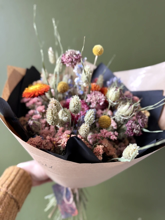 a dried flower bouquet in bright colours wrapped in black tissue and kraft paper held by a person in a mustard coloured jumper with their left arm against a green wall