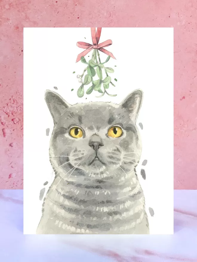 A Christmas card featuring a hand painted design of a Grey British Shorthair Cat, stood upright on a marble surface.