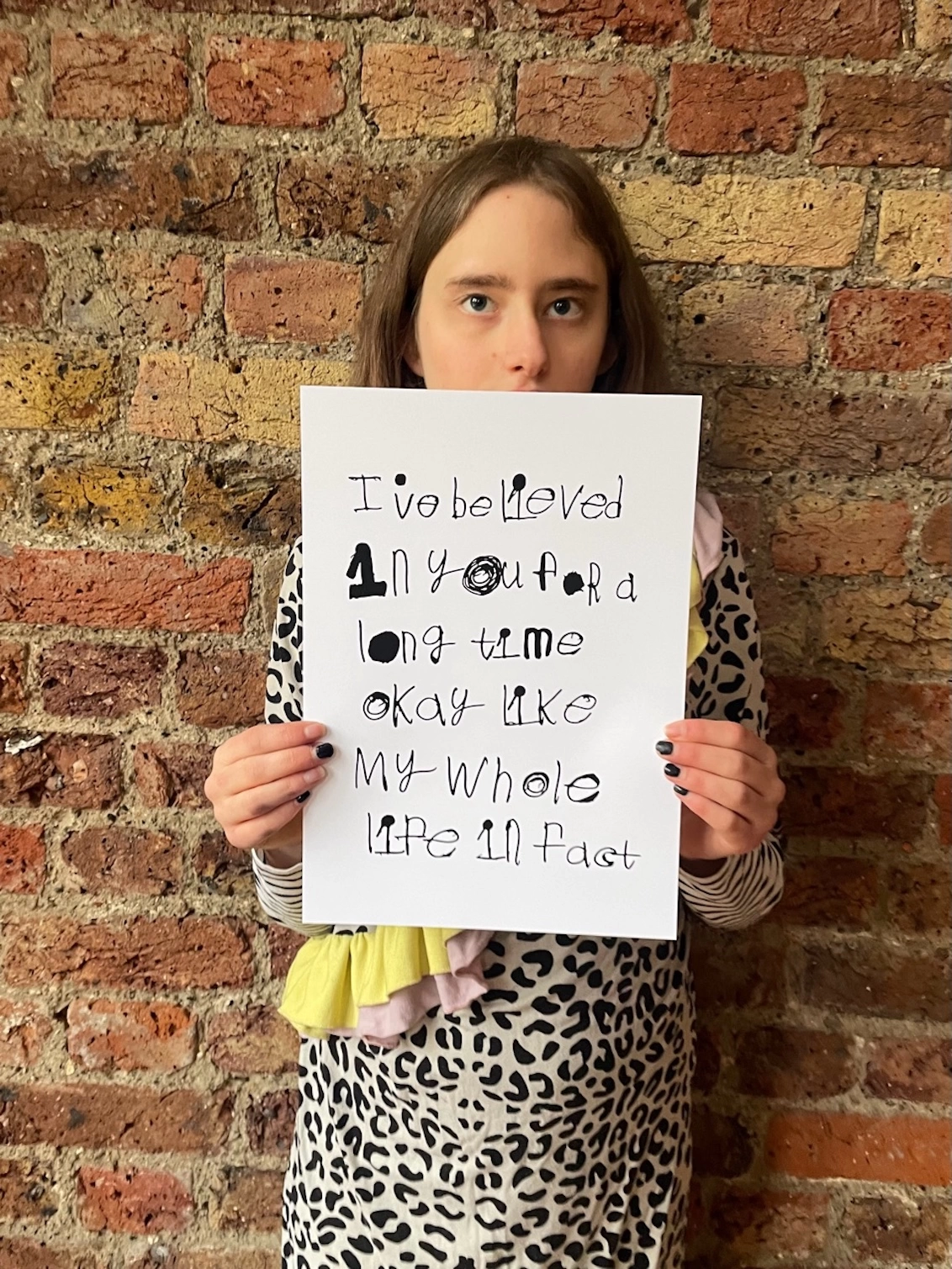 Piper a deaf, teenage, autistic artist from Hackney hold her a4 artwork up in front of a brick wall. The art is black handwritten unique font on a white background.