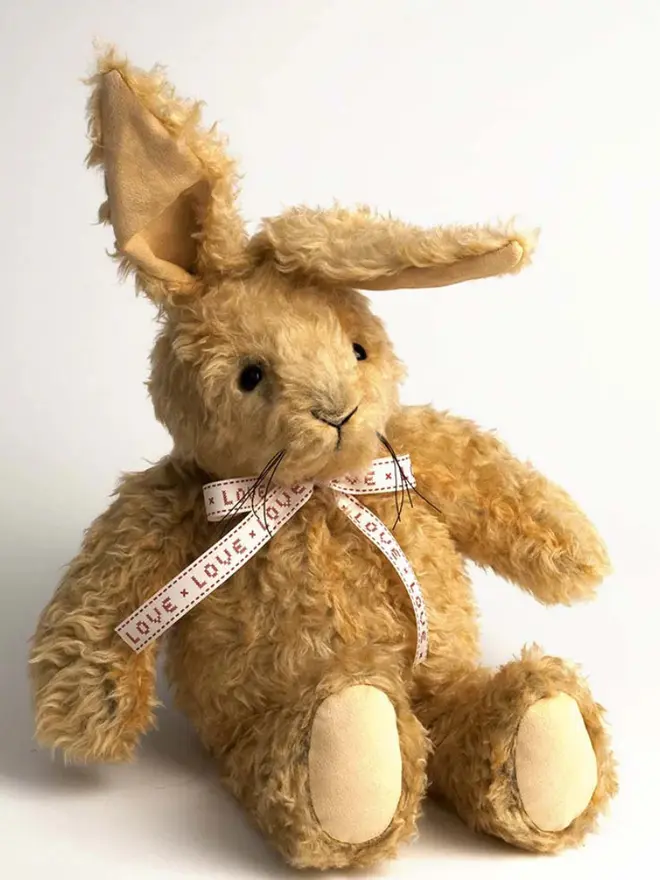 Handmade brown stuffed rabbit with long ears and a pink ribbon bow sitting upright against a plain background.