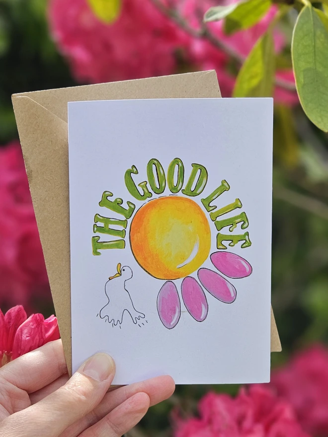 A white card with an illustration of a pink flower and a white bird with The Good Life written using a retro style font in green. 