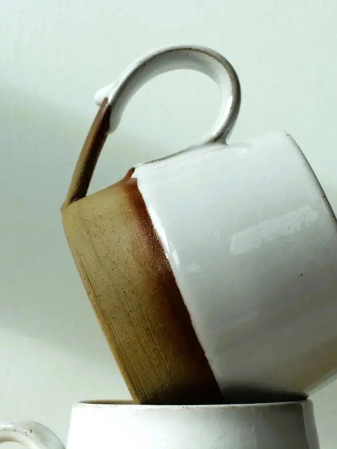 Handmade ceramic mug with a glossy white upper glaze and a natural toasted clay base, against a soft green wall background.