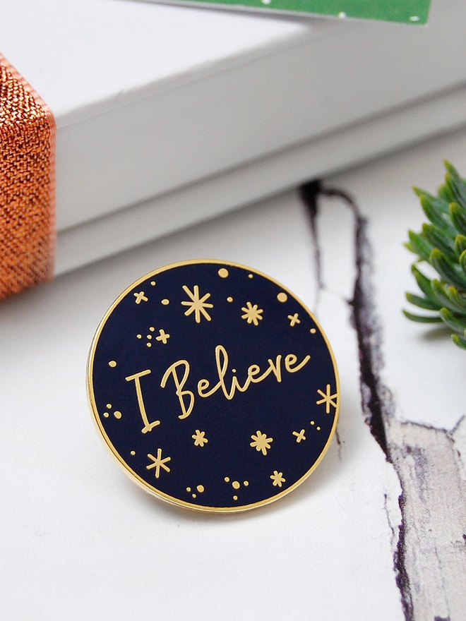A navy blue and gold enamel pin badge with a starry design and the words "I Believe" is resting on a white wooden desk.
